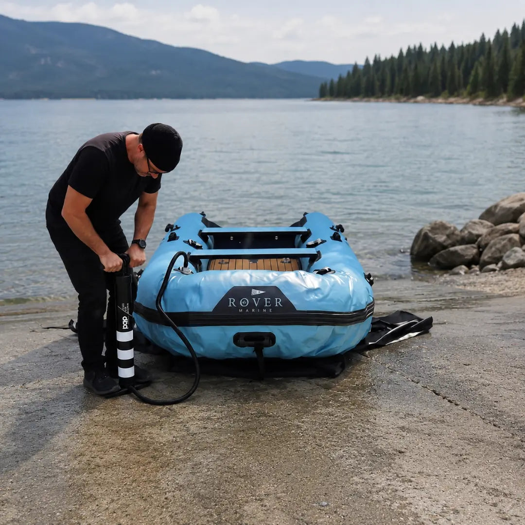 Person inflating a blue inflatable boat on a lakeside with mountains in the background